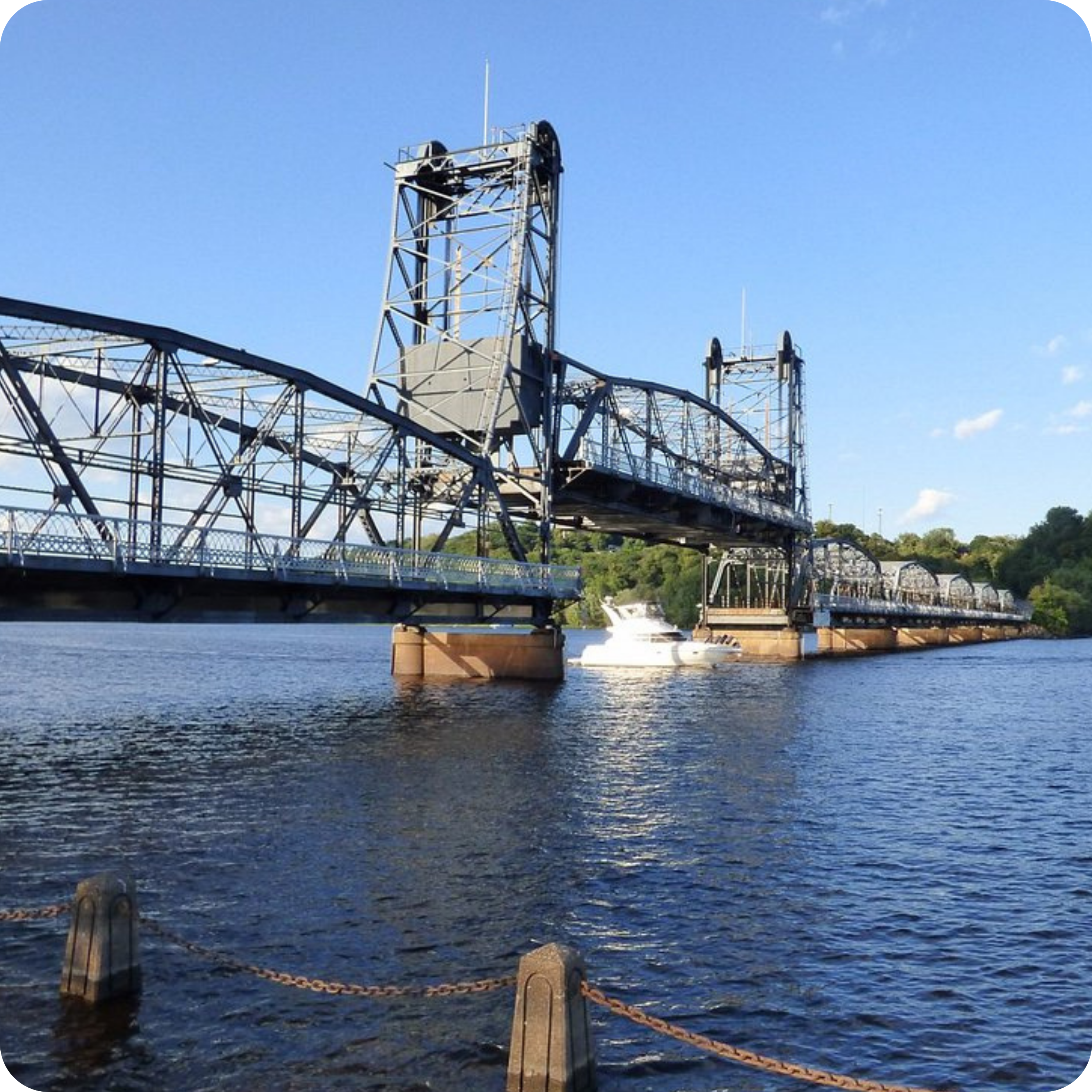 A metal truss bridge spans a calm body of water in Stillwater, Minnesota, with a small white boat beneath it and trees in the background.